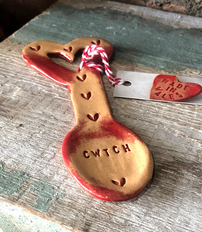Ceramic Welsh lovespoon in a red glaze decorated with hearts and the word cwtch. Has a printed label saying Made in Wales.