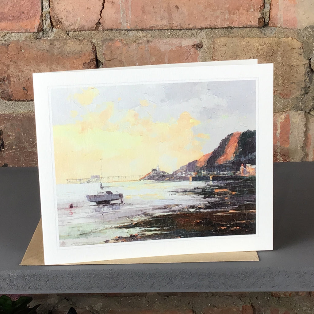 Greetings card of Mumbles Bay at first light with an illuminated cloudy sky and boat in the foreground.